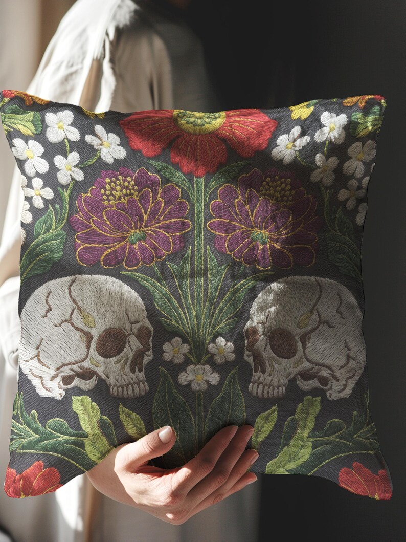 Grimoire and Goblet gothic pillow cover featuring mirrored ivory human skulls surrounded by detailed floral and botanical elements including deep red flowers, purple blooms with gold outlining, small white blossoms, and layered green foliage, arranged in a symmetrical composition on a charcoal background, designed with a high-detail faux embroidery print that mimics stitched texture while remaining smooth, creating a dark academia and gothic botanical aesthetic decorative cushion cover