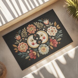 Witchy outdoor doormat featuring a central all-seeing eye, crescent moons, and floral details in an embroidery-style design. This weatherproof welcome mat measures 26x18 inches and is ideal for porches or patio entries. Symbolic celestial motifs offer intuitive protection and spiritual elegance for magical home decor.