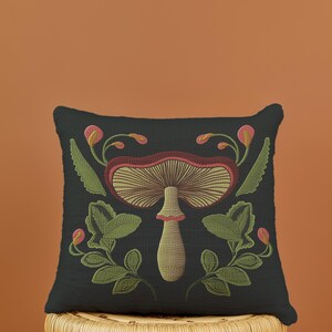 A dark woven pillow with a detailed embroidered mushroom and botanical design in deep red, green, and gold tones. Made with a cotton-poly blend.