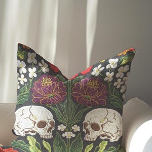 Grimoire and Goblet gothic pillow cover featuring mirrored ivory human skulls surrounded by detailed floral and botanical elements including deep red flowers, purple blooms with gold outlining, small white blossoms, and layered green foliage, arranged in a symmetrical composition on a charcoal background, designed with a high-detail faux embroidery print that mimics stitched texture while remaining smooth, creating a dark academia and gothic botanical aesthetic decorative cushion cover