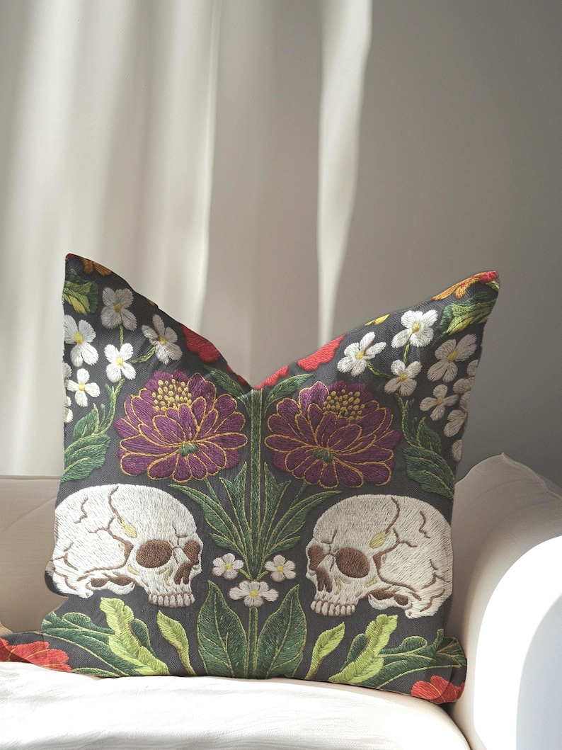 Grimoire and Goblet gothic pillow cover featuring mirrored ivory human skulls surrounded by detailed floral and botanical elements including deep red flowers, purple blooms with gold outlining, small white blossoms, and layered green foliage, arranged in a symmetrical composition on a charcoal background, designed with a high-detail faux embroidery print that mimics stitched texture while remaining smooth, creating a dark academia and gothic botanical aesthetic decorative cushion cover