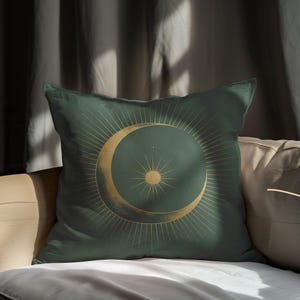 Puede incluir: Una almohada verde con un diseño de luna creciente y sol dorado. La almohada tiene una textura sutil y una estética minimalista.