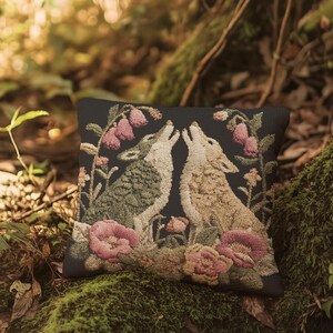 A square woven tapestry pillow with two howling wolves on a black background. The wolves are surrounded by pink and green botanical flowers, creating a vintage, folkloric scene. This decorative cushion is perfect for cottagecore, dark academia, and gothic home decor.