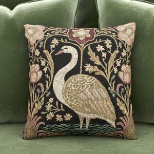 Könnte beinhalten: Quadratisches Zierkissen mit schwarzem Hintergrund und Schwanenmotiv. Der Schwan ist cremefarben mit einem detaillierten Muster. Das Kissen hat einen floralen Rand mit rosa und goldenen Akzenten, vor einem grünen Hintergrund.