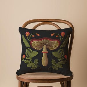 A dark woven pillow with a detailed embroidered mushroom and botanical design in deep red, green, and gold tones. Made with a cotton-poly blend.