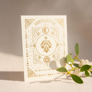 Puede incluir: Una tarjeta crema con detalles dorados que presenta la palabra "MISTLETOE" y diseños celestiales decorativos. Una ramita de muérdago con hojas verdes y bayas blancas descansa junto a la tarjeta. El texto "Love, Protection & New Beginnings" también es visible.