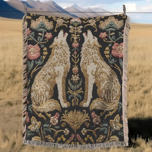 May include: A woven throw blanket with a black background, featuring two howling wolves in beige and gold, surrounded by floral patterns in pink, green, and gold. The blanket has a fringed edge and is hanging on a clothesline.