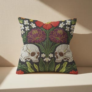 Grimoire and Goblet gothic pillow cover featuring mirrored ivory human skulls surrounded by detailed floral and botanical elements including deep red flowers, purple blooms with gold outlining, small white blossoms, and layered green foliage, arranged in a symmetrical composition on a charcoal background, designed with a high-detail faux embroidery print that mimics stitched texture while remaining smooth, creating a dark academia and gothic botanical aesthetic decorative cushion cover
