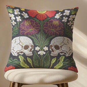 Grimoire and Goblet gothic pillow cover featuring mirrored ivory human skulls surrounded by detailed floral and botanical elements including deep red flowers, purple blooms with gold outlining, small white blossoms, and layered green foliage, arranged in a symmetrical composition on a charcoal background, designed with a high-detail faux embroidery print that mimics stitched texture while remaining smooth, creating a dark academia and gothic botanical aesthetic decorative cushion cover