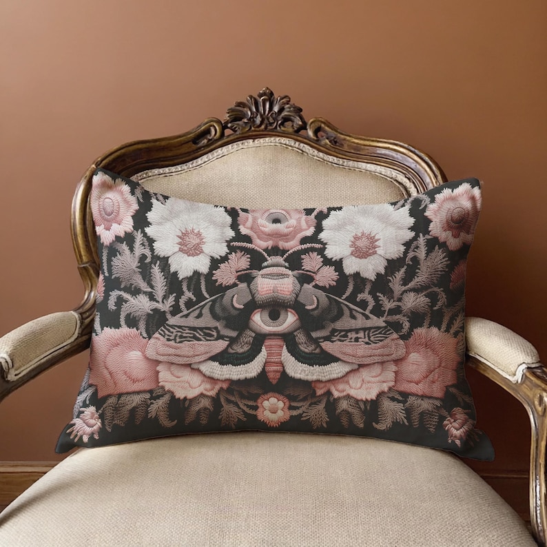 Gothic moth eye botanical lumbar pillow with mystical embroidered wings on dark fabric. Cottagecore throw pillow featuring protective moth symbol surrounded by pink roses, white wildflowers & sacred botanicals. Witchy home decor with medieval tapestry aesthetic. Perfect for dark academia spaces, bohemian decor & occult art lovers. 20x12 lumbar size.