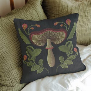 A dark woven pillow with a detailed embroidered mushroom and botanical design in deep red, green, and gold tones. Made with a cotton-poly blend.