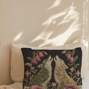 A square woven tapestry pillow with two howling wolves on a black background. The wolves are surrounded by pink and green botanical flowers, creating a vintage, folkloric scene. This decorative cushion is perfect for cottagecore, dark academia, and gothic home decor.
