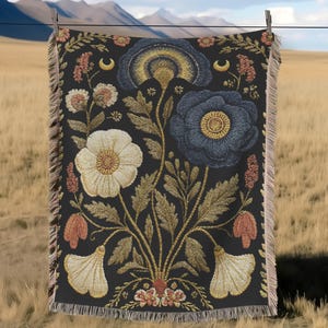 May include: A black woven throw blanket with a floral design, featuring large white, blue, and gold flowers. The blanket has a fringed edge and is hanging on a clothesline against a backdrop of mountains and a field.