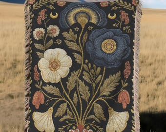 Può includere: Una coperta tessuta nera con un motivo floreale, con grandi fiori bianchi, blu e oro. La coperta ha un bordo con frange ed è appesa a un filo per stendere i panni sullo sfondo di montagne e un campo.