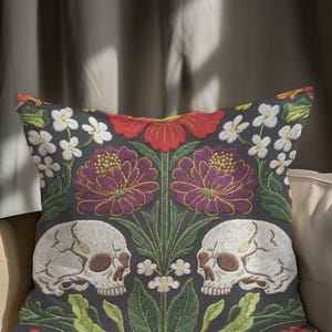 Grimoire and Goblet gothic pillow cover featuring mirrored ivory human skulls surrounded by detailed floral and botanical elements including deep red flowers, purple blooms with gold outlining, small white blossoms, and layered green foliage, arranged in a symmetrical composition on a charcoal background, designed with a high-detail faux embroidery print that mimics stitched texture while remaining smooth, creating a dark academia and gothic botanical aesthetic decorative cushion cover
