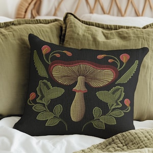 A dark woven pillow with a detailed embroidered mushroom and botanical design in deep red, green, and gold tones. Made with a cotton-poly blend.