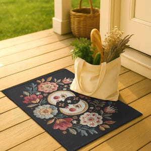 Witchy outdoor doormat featuring a central all-seeing eye, crescent moons, and floral details in an embroidery-style design. This weatherproof welcome mat measures 26x18 inches and is ideal for porches or patio entries. Symbolic celestial motifs offer intuitive protection and spiritual elegance for magical home decor.
