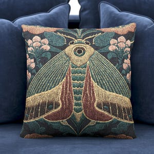 Puede incluir: Cojín cuadrado decorativo con un diseño detallado de polilla en tonos turquesa, dorado y burdeos, sobre un fondo oscuro con detalles florales. El cojín está colocado sobre un sofá azul marino, creando un contraste llamativo.