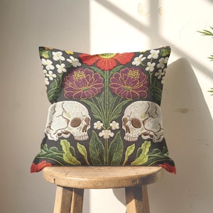 Grimoire and Goblet gothic pillow cover featuring mirrored ivory human skulls surrounded by detailed floral and botanical elements including deep red flowers, purple blooms with gold outlining, small white blossoms, and layered green foliage, arranged in a symmetrical composition on a charcoal background, designed with a high-detail faux embroidery print that mimics stitched texture while remaining smooth, creating a dark academia and gothic botanical aesthetic decorative cushion cover