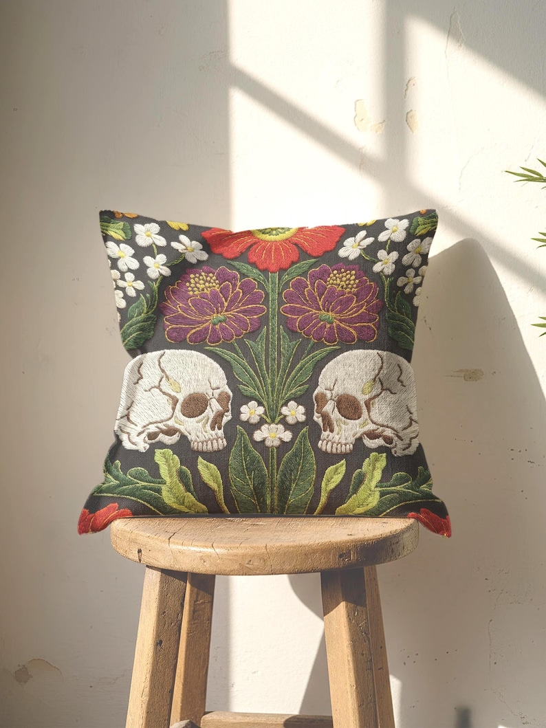 Grimoire and Goblet gothic pillow cover featuring mirrored ivory human skulls surrounded by detailed floral and botanical elements including deep red flowers, purple blooms with gold outlining, small white blossoms, and layered green foliage, arranged in a symmetrical composition on a charcoal background, designed with a high-detail faux embroidery print that mimics stitched texture while remaining smooth, creating a dark academia and gothic botanical aesthetic decorative cushion cover