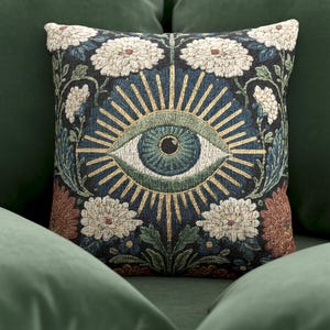 May include: Square decorative pillow with an embroidered eye design. The pillow features a central eye motif with radiating gold lines, surrounded by white floral and botanical patterns on a dark background. The pillow is placed on a green couch.
