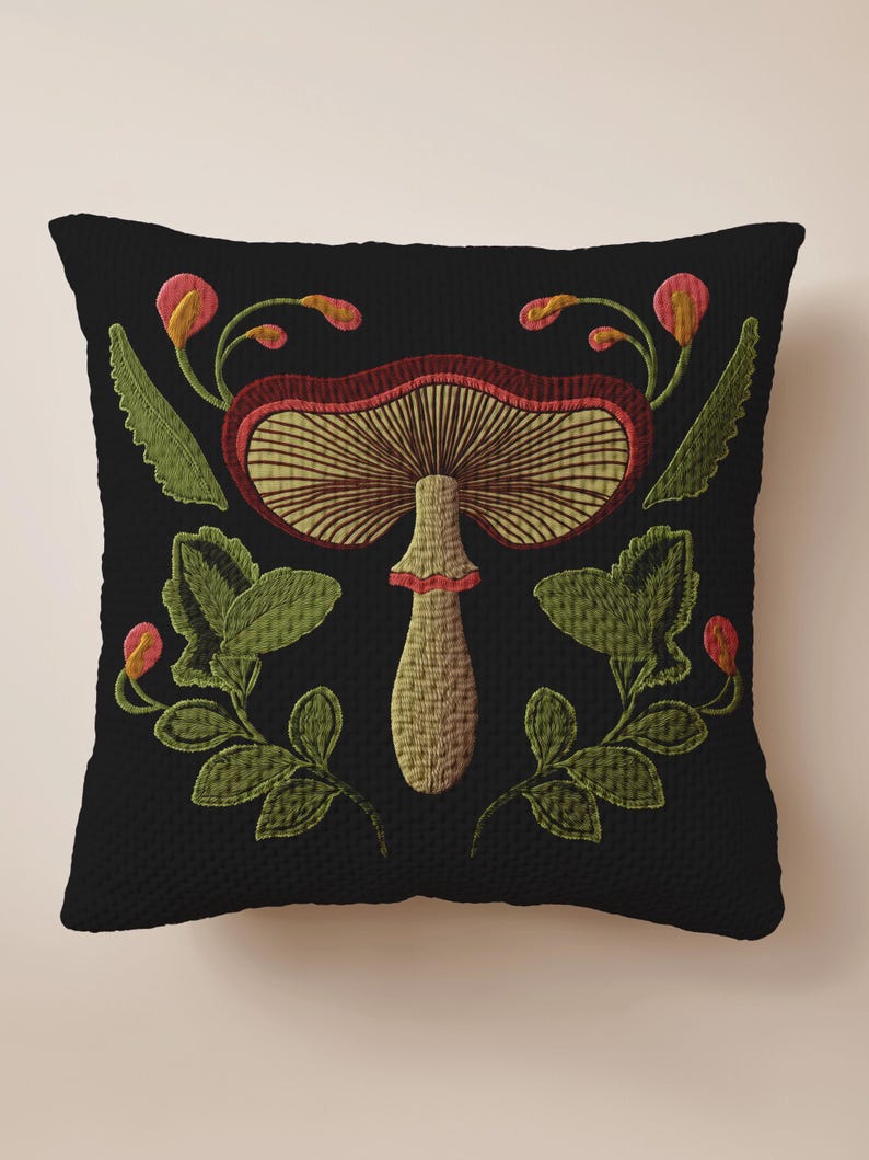 A dark woven pillow with a detailed embroidered mushroom and botanical design in deep red, green, and gold tones. Made with a cotton-poly blend.
