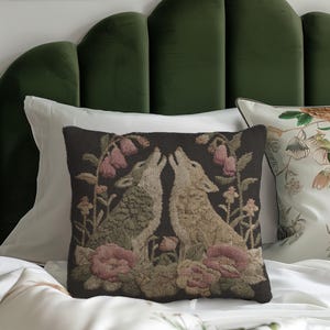 A square woven tapestry pillow with two howling wolves on a black background. The wolves are surrounded by pink and green botanical flowers, creating a vintage, folkloric scene. This decorative cushion is perfect for cottagecore, dark academia, and gothic home decor.