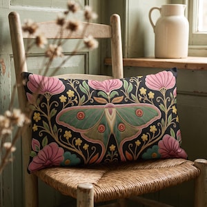 May include: A rectangular throw pillow featuring a black background with a central green moth illustration surrounded by symmetrical pink flowers, yellow blossoms, and teal accents. The design is intricate with detailed embroidery-style patterns.