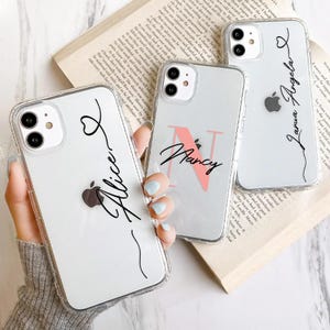 Puede incluir: Tres fundas transparentes para teléfono con nombres personalizados y un diseño de corazón. Los nombres son "Alice", "Nancy" y "Laura Angela".
