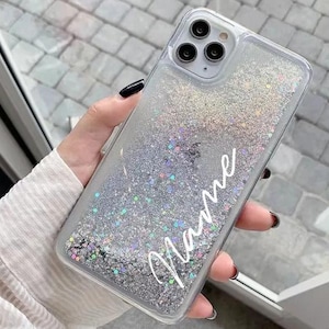 Peut inclure: Une coque de téléphone transparente avec un motif de paillettes argentées et le mot "Name" écrit en blanc. La coque est conçue pour un téléphone avec un renflement de caméra.