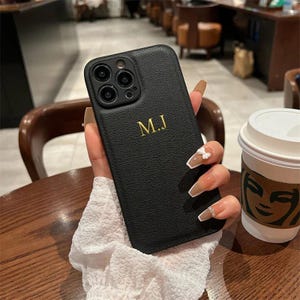 Peut inclure: Étui de téléphone en cuir noir avec un monogramme doré "M.J." et une découpe pour l'objectif de l'appareil photo.