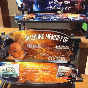 May include: Memorial plaques with the text "IN LOVING MEMORY OF" and names, dates, and photos. The plaques have a curved top and are supported by black stands. The background features a celestial design.