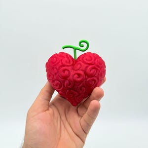 May include: A red, heart-shaped object with a green stem and a swirled design. The object is held in a hand against a white background. The object appears to be a prop or a decorative item.