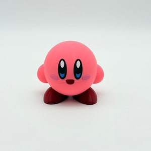 May include: A pink, spherical character with small arms, rosy cheeks, and large, oval eyes. The character has dark red feet and a small, smiling mouth. This 3D-printed figure is a popular video game character.