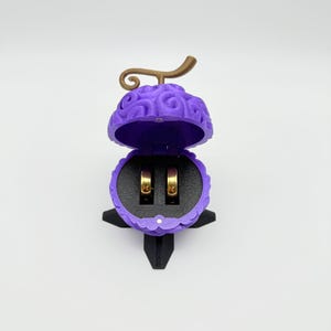 Könnte beinhalten: Eine offene, lila Ringbox in Fruchtform mit einem goldfarbenen Stiel und zwei goldenen Ringen im Inneren. Die Box steht auf einem schwarzen Sockel, und der Innenraum ist schwarz. Die Box ist als Teufelsfrucht gestaltet.