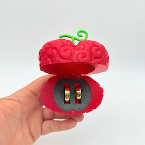 Könnte beinhalten: Eine rote, fruchtförmige Ringbox mit einem grünen Stiel, die geöffnet ist, um zwei goldene Ringe zu enthüllen. Die Box hat eine strukturierte Oberfläche und einen dunkelgrauen Innenraum. Die Ringe werden vor einem grauen Hintergrund präsentiert.