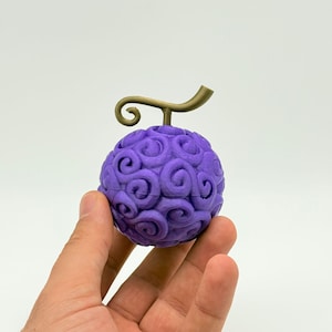 May include: A purple, spherical fruit with a swirling pattern and a gold-colored stem. The fruit is held in a hand against a white background. The design is reminiscent of a fictional item.