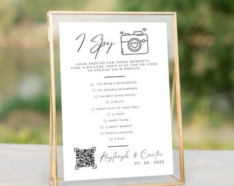 May include: A gold framed sign with the text "I Spy" and a camera graphic. The sign includes a list of photo prompts and a QR code. The names "Kayleigh & Carter" and the date "07.26.2025" are at the bottom.