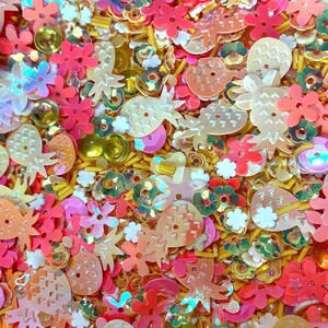 May include: A vibrant mix of craft supplies, including pineapple and flower-shaped sequins in shades of pink, orange, and iridescent white. Gold and yellow accents add sparkle to this colorful assortment, perfect for crafting projects.