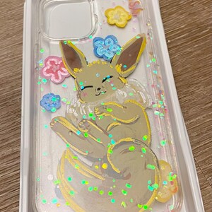 May include: A clear phone case with a cartoon illustration of a brown and yellow Eevee with pink, blue, and yellow flowers. The case is filled with iridescent glitter.