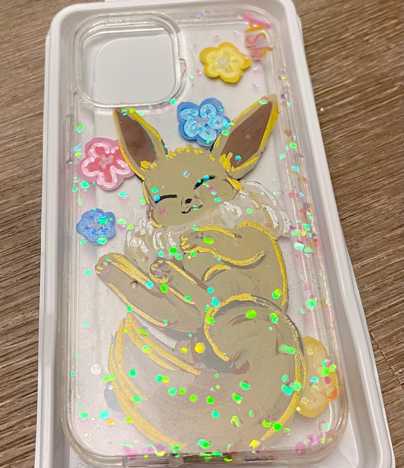 May include: A clear phone case with a cartoon illustration of a brown and yellow Eevee with pink, blue, and yellow flowers. The case is filled with iridescent glitter.