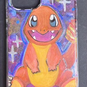 May include: A clear phone case with a hand-painted design of a cartoon orange and yellow Pokemon character, Charmander, sitting on a Pokeball. The background is a blue and purple galaxy with white stars.