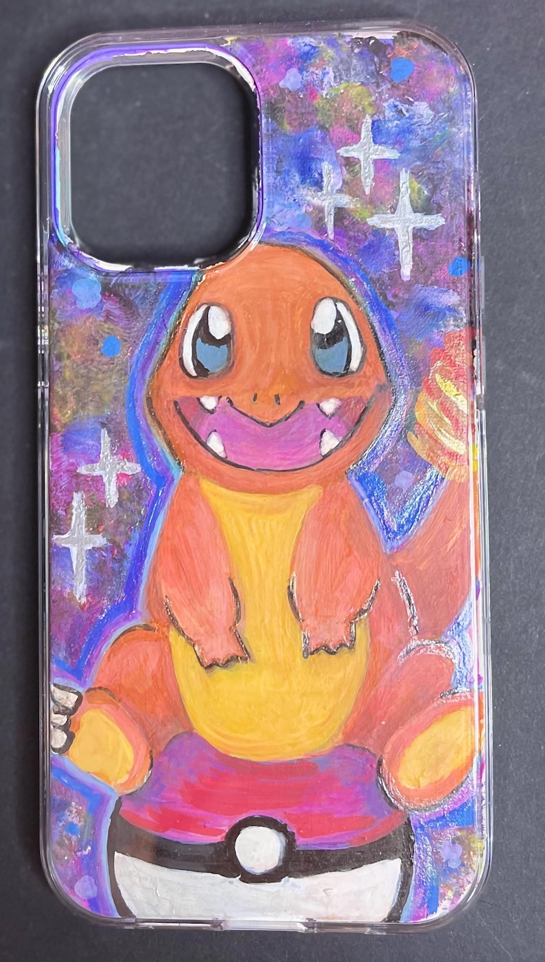 May include: A clear phone case with a hand-painted design of a cartoon orange and yellow Pokemon character, Charmander, sitting on a Pokeball. The background is a blue and purple galaxy with white stars.