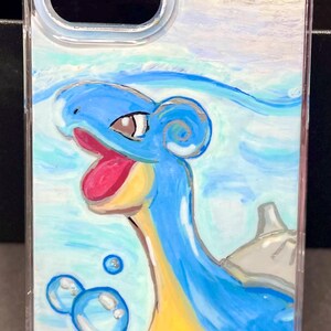 May include: A clear phone case with a hand-painted design of a blue and yellow Pokemon character, with the name "Ash" written in pink at the top.