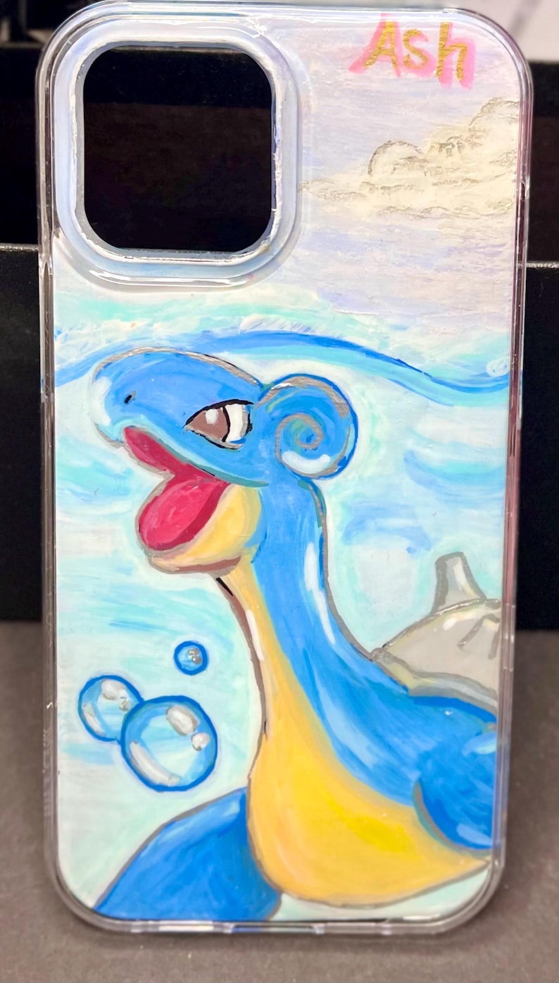 May include: A clear phone case with a hand-painted design of a blue and yellow Pokemon character, with the name "Ash" written in pink at the top.