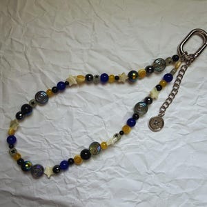 May include: A beaded keychain with a silver clasp and chain. The beads are a mix of blue, yellow, black, and iridescent colors, with star-shaped and circular accents. A small, round charm hangs from the chain.