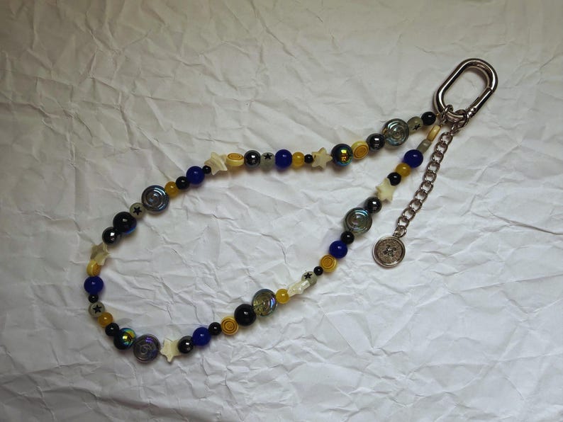 May include: A beaded keychain with a silver clasp and chain. The beads are a mix of blue, yellow, black, and iridescent colors, with star-shaped and circular accents. A small, round charm hangs from the chain.