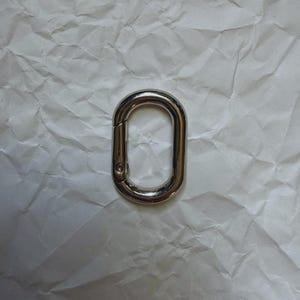May include: A shiny, silver-colored oval carabiner clip. The clip is made of metal and has a spring-loaded gate for secure closure. The background is a crumpled white paper.