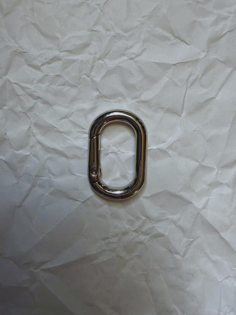 May include: A shiny, silver-colored oval carabiner clip. The clip is made of metal and has a spring-loaded gate for secure closure. The background is a crumpled white paper.