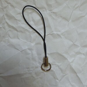 May include: A black lanyard with a gold-colored metal ring and barrel. The lanyard is looped and attached to the metal barrel. The background is a crumpled white paper.